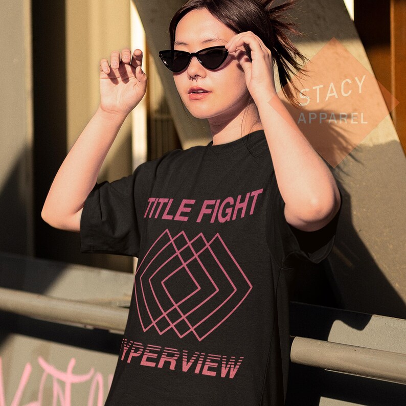 Puede incluir: Camiseta negra con las palabras "TITLE FIGHT" en rosa, encima de un dise&ntilde;o geom&eacute;trico rosa, y la palabra "HYPERVIEW" debajo. La camiseta la lleva una persona con gafas de sol negras.