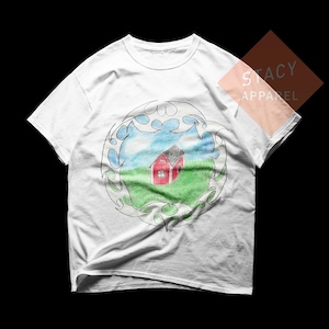 May include: White t-shirt featuring a colorful drawing of a red house with a gray roof, set in a green field under a blue sky. The artwork is framed by a decorative, swirling border. The shirt has the text "STACY APPAREL".