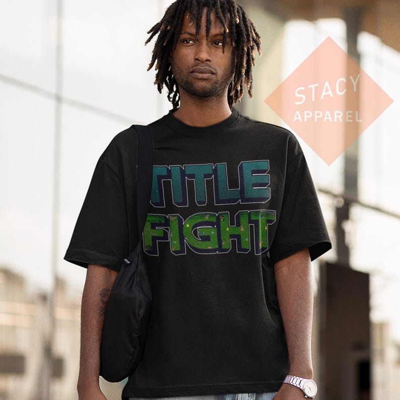 K&ouml;nnte beinhalten: Schwarzes T-Shirt mit dem Schriftzug "TITLE FIGHT" in einem teal- und gr&uuml;nen 3D-Design. Das kurz&auml;rmelige Shirt hat einen Rundhalsausschnitt. Der Markenname "STACY APPAREL" befindet sich in einem pfirsichfarbenen Diamanten oben rechts.