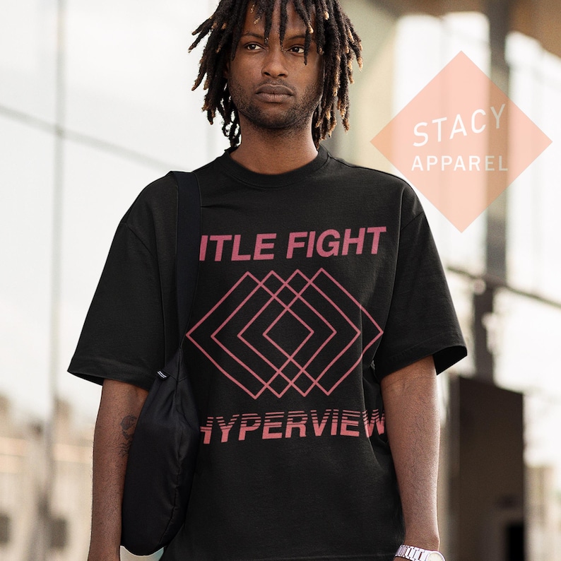 Puede incluir: Camiseta negra con las palabras rosas "ITLE FIGHT", sobre un dise&ntilde;o geom&eacute;trico rosa y la palabra "HYPERVIEW". La camiseta es de la colecci&oacute;n Stacy Apparel.
