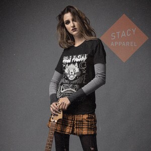 May include: Black t-shirt with a cat graphic and the text "Wile Fight". The shirt is layered over a striped long-sleeve shirt. The person is wearing a plaid skirt and holding a guitar. The Stacy Apparel logo is in the background.