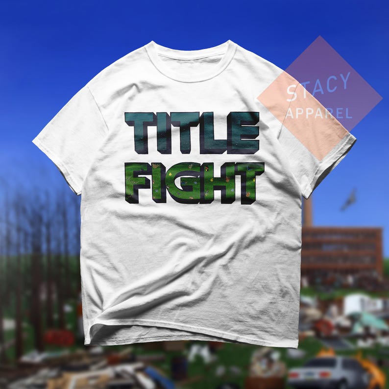 K&ouml;nnte beinhalten: Wei&szlig;es T-Shirt mit dem Schriftzug "TITLE FIGHT" in einem 3D-Design in Gr&uuml;n und T&uuml;rkis. Das Shirt besteht aus einem weichen Material und eignet sich f&uuml;r Freizeitkleidung. Das Stacy Apparel-Logo ist oben rechts sichtbar.