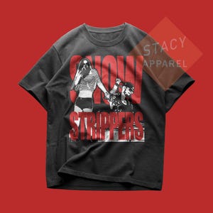 May include: Black t-shirt with a graphic design featuring two figures and the words "SNOW STRIPPERS" in red. The shirt has a crew neck and short sleeves. The Stacy Apparel logo is visible.