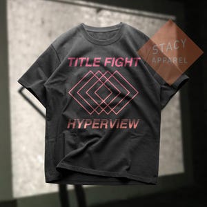 Puede incluir: Camiseta gris oscuro con las palabras "TITLE FIGHT" y "HYPERVIEW" en rosa, junto con un dise&ntilde;o geom&eacute;trico. La camiseta tiene cuello redondo y mangas cortas. El logotipo de Stacy Apparel es visible en la esquina superior derecha.