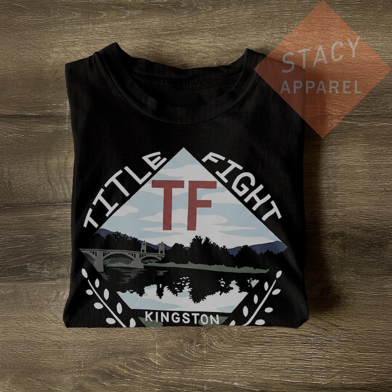 Puede incluir: Camiseta negra con un gr&aacute;fico que muestra un puente sobre el agua, monta&ntilde;as y el texto "TITLE FIGHT KINGSTON". Las letras "TF" son rojas. La camiseta est&aacute; doblada.