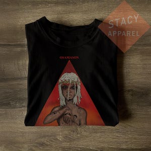Puede incluir: Camiseta negra con un gráfico de una persona con rastas blancas y una corona de espinas, sobre un triángulo rojo. La palabra "OSAMASON" está impresa encima del gráfico. La camiseta es de Stacy Apparel.