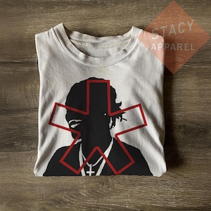 Könnte beinhalten: Ein gefaltetes, hellgraues T-Shirt mit einer schwarzen Silhouette einer Person und einem roten sternartigen Design um Kopf und Schultern. Das Shirt hat einen Rundhalsausschnitt und ist aus weichem Material. Der Text "STACY APPAREL" ist rechts oben sichtbar.