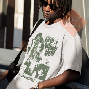 Puede incluir: Camiseta gris claro con un dise&ntilde;o gr&aacute;fico verde y blanco. El dise&ntilde;o incluye las palabras "Bea Bajoo Bee" y "beatopia", as&iacute; como ilustraciones de mujeres y estrellas. La camiseta es de manga corta.