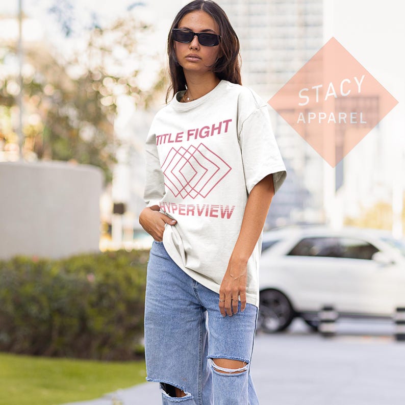 Puede incluir: Una camiseta blanca con las palabras "TITLE FIGHT" y "HYPERVIEW" en una fuente rosa-roja. La camiseta presenta un dise&ntilde;o geom&eacute;trico en el centro. La modelo lleva vaqueros azules y gafas de sol. El logotipo de Stacy Apparel est&aacute; en el fondo.