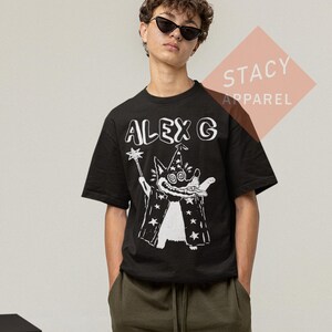 Puede incluir: Camiseta negra con un gr&aacute;fico blanco de un mago de dibujos animados sosteniendo una varita y usando un sombrero y una capa cubiertos de estrellas. El texto "ALEX G" est&aacute; encima del gr&aacute;fico. La camiseta es de la colecci&oacute;n Stacy Apparel.