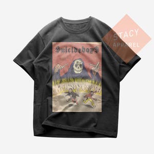 Op de afbeelding: Donkergrijs t-shirt met een grafische print. De print toont een figuur met een schedelgezicht met de tekst "Suicideboys" erboven, en twee figuren die wegrennen van de vlammen. Het shirt is van Stacy Apparel.