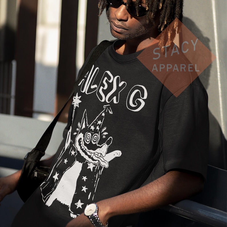 Puede incluir: Camiseta negra de cuello redondo con el texto blanco "ALEX G" y una ilustraci&oacute;n de un perro mago de dibujos animados. El perro lleva un sombrero y una capa de mago. El logotipo de Stacy Apparel es visible en la esquina superior derecha.