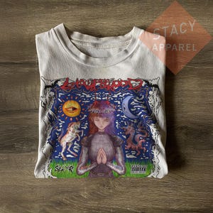 May include: White t-shirt with a colorful graphic design. The design features a woman with red hair, a sun, moon, and a dragon. The text "Stay Blood" is at the top, and "Slayr" is at the bottom. The shirt is folded on a wooden surface.