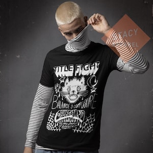 May include: Black t-shirt featuring a white cat graphic, the words "TITLE FIGHT", "BALANCE & COMPOSURE", and "CHUGGAARD". The t-shirt is layered over a striped long-sleeved top.
