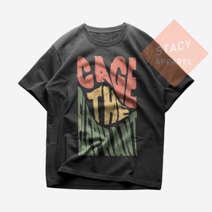 Könnte beinhalten: Dunkelgraues T-Shirt mit dem Schriftzug "CAGE THE ELEPHANT" in einem farbenfrohen Retro-Schriftzug. Der Text befindet sich in einem vertikalen Rechteck, wobei der Name der Band in Rot, Gelb und Grün gehalten ist. Das Shirt hat kurze Ärmel und einen Rundhalsausschnitt.