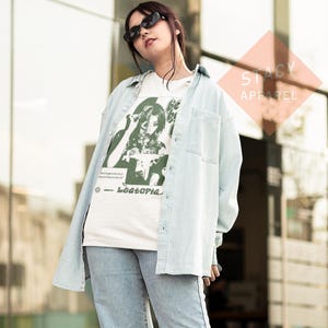 Puede incluir: Una camisa vaquera azul claro sobre una camiseta gr&aacute;fica blanca y vaqueros azul claro. La camiseta presenta un gr&aacute;fico verde y blanco con el texto "Beatopia". La camisa tiene dos bolsillos en el pecho.