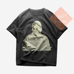 Peut inclure: T-shirt gris foncé avec une image délavée d'un visage et du haut du corps d'une personne. Le texte "DEAN BLUNT" est imprimé dans une police vieillie sous l'image. Le t-shirt est de Stacy Apparel.
