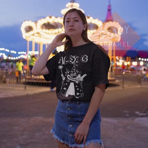 Puede incluir: Camiseta negra con el texto "ALEX G" y un dise&ntilde;o de mago de dibujos animados en blanco. El mago lleva una t&uacute;nica cubierta de estrellas y sostiene una varita. La modelo lleva una falda vaquera.