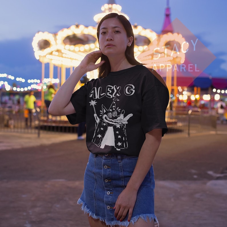 Puede incluir: Camiseta negra con el texto "ALEX G" y un dise&ntilde;o de mago de dibujos animados en blanco. El mago lleva una t&uacute;nica cubierta de estrellas y sostiene una varita. La modelo lleva una falda vaquera.
