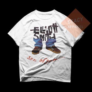 Puede incluir: Camiseta blanca con un gr&aacute;fico de piernas con vaqueros azules y zapatos marrones, con el nombre "ELLIOTT SMITH" en letras negras. Las palabras "Son of Sam" est&aacute;n escritas en rojo debajo.