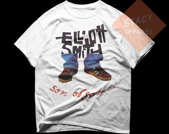May include: White t-shirt featuring a graphic of legs in blue jeans and brown shoes, with the name "ELLIOTT SMITH" in black block letters. The words "Son of Sam" are written in red script below the graphic.