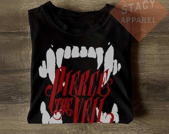 Pode incluir: Camiseta preta com um desenho de presas brancas e as palavras "Pierce the Veil" em vermelho. A camiseta é da Stacy Apparel. O desenho está centrado na frente da camiseta dobrada.