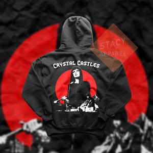 May include: Black hoodie featuring a graphic of a woman in front of a red circle, with the text "CRYSTAL CASTLES" above. The design is in white. The hoodie has a hood and is made of a soft material. "STACY APPAREL" is printed on the upper right.