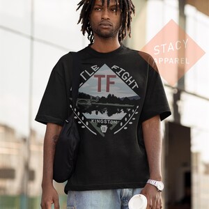 Puede incluir: Camiseta negra con un dise&ntilde;o gr&aacute;fico que presenta el texto "TF FIGHT" y "KINGSTON 13". El dise&ntilde;o incluye un paisaje con un puente y agua. La camiseta la lleva una persona.