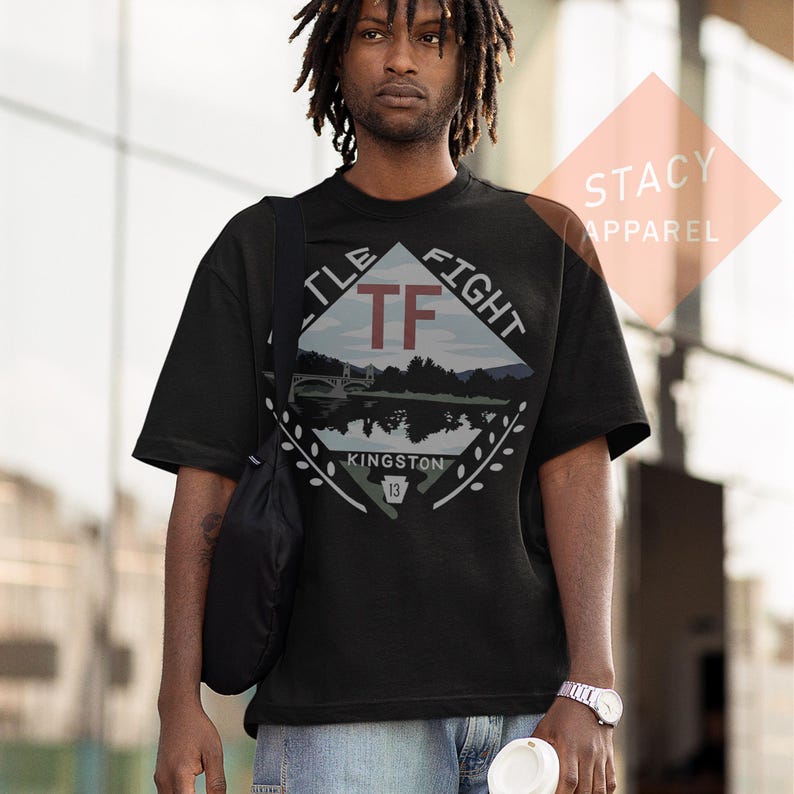 Puede incluir: Camiseta negra con un dise&ntilde;o gr&aacute;fico que presenta el texto "TF FIGHT" y "KINGSTON 13". El dise&ntilde;o incluye un paisaje con un puente y agua. La camiseta la lleva una persona.