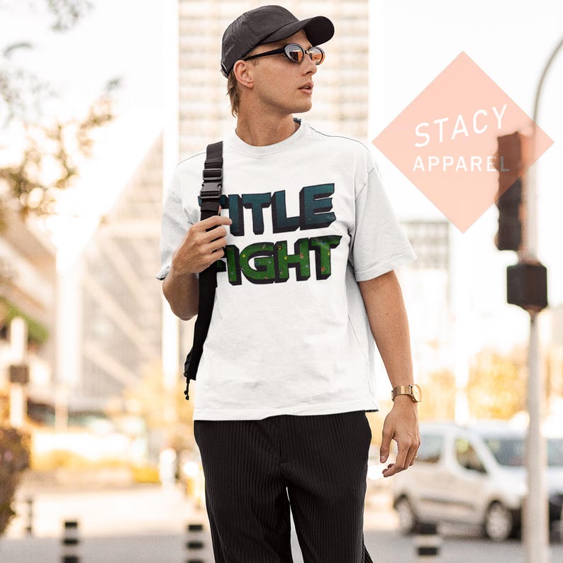 K&ouml;nnte beinhalten: Wei&szlig;es T-Shirt mit dem Schriftzug "TITLE FIGHT" in 3D-Optik, mit blauen und gr&uuml;nen Farben. Das T-Shirt wird von einer Person getragen, die eine schwarze Kappe und Sonnenbrille tr&auml;gt. Das T-Shirt ist von Stacy Apparel.