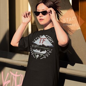 Puede incluir: Camiseta negra con un dise&ntilde;o gr&aacute;fico que presenta el texto "TITLE FIGHT" y "TF" en forma de diamante. El dise&ntilde;o incluye un paisaje con un puente y la palabra "KINGSTON". La camiseta es de "STACY APPAREL".