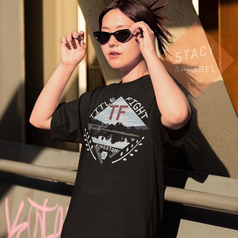 Puede incluir: Camiseta negra con un dise&ntilde;o gr&aacute;fico que presenta el texto "TITLE FIGHT" y "TF" en forma de diamante. El dise&ntilde;o incluye un paisaje con un puente y la palabra "KINGSTON". La camiseta es de "STACY APPAREL".
