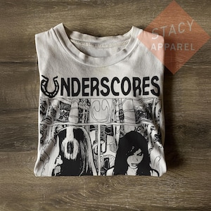 Puede incluir: Una camiseta blanca con la palabra "UNDERSCORES" en letras negras. La camiseta presenta un gráfico en blanco y negro de dos figuras. La camiseta está doblada sobre una superficie de madera.