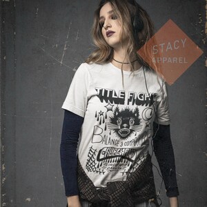May include: A white t-shirt featuring a black cat graphic, stars, and the words "TITLE FIGHT", "BALANCE 3 COMPOSED", and "CHUGGER". The shirt is layered over a navy blue long-sleeved top and a plaid shirt tied at the waist. The Stacy Apparel logo is visible.