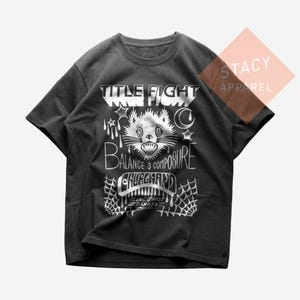 May include: Dark grey t-shirt featuring a white graphic design. The design includes the words "TITLE FIGHT", a cat illustration, and the words "BALANCE & COMPOSURE". The shirt is from Stacy Apparel.