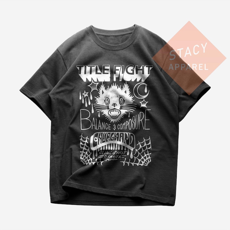 May include: Dark grey t-shirt featuring a white graphic design. The design includes the words "TITLE FIGHT", a cat illustration, and the words "BALANCE & COMPOSURE". The shirt is from Stacy Apparel.