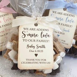 May include: Three clear plastic favor bags filled with s'mores ingredients, each tied with a ribbon and a rectangular paper tag. The tags feature the text "WE ARE ADDING S'MORE LOVE TO OUR FAMILY!" along with a name, a due date, and a thank you message.