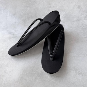 Puede incluir: Un par de sandalias geta negras, calzado tradicional japonés. Cuentan con una parte superior de tela texturizada y una correa tipo tanga. Las sandalias tienen una plataforma elevada y una suela blanca.