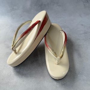 Puede incluir: Un par de sandalias geta japonesas de color crema con suela de plataforma. Las correas son doradas y rojas. Las sandalias están hechas de un material brillante y están sobre un fondo gris.