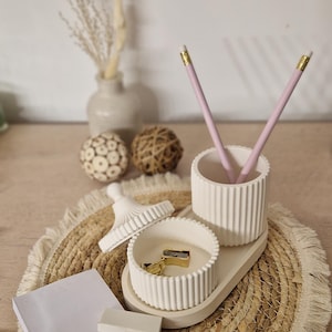 Può includere: Un set di organizzazione da scrivania con un portapenne bianco, un piccolo vassoio e un coperchio abbinato. Il set include due matite rosa, un temperamatite dorato e un blocco note. L'organizer è posizionato su un sottobicchiere intrecciato.