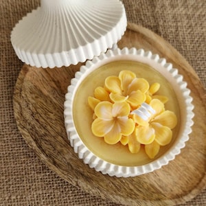 Peut inclure: Un récipient rond blanc avec un couvercle au design cannelé. À l'intérieur se trouve une surface en cire jaune lisse surmontée d'un groupe de fleurs jaunes en trois dimensions avec des centres blancs et un petit élément décoratif blanc.