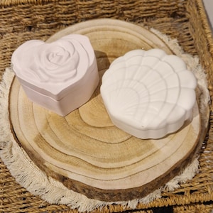 Könnte beinhalten: Zwei dekorative Schmuckdosen in Weiß. Eine ist herzförmig mit Rosenmuster, die andere hat die Form einer Muschel. Beide liegen auf einer Holzscheibe mit einem geflochtenen Korbhintergrund.