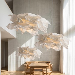 May include: Three large, cloud-like pendant lights made of white, layered paper hang above a wooden dining table and chairs. The room has a modern design with concrete walls, hardwood floors, and a large window with white curtains.