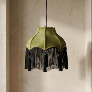Puede incluir: Pantalla de lámpara colgante con exterior de terciopelo verde y flecos negros. La pantalla tiene un borde festoneado y un ribete decorativo. Los flecos negros cuelgan del borde inferior.