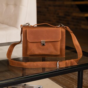 May include: A brown leather messenger bag with a flap closure and adjustable shoulder strap. The bag features a front pocket with a silver-colored clasp. The bag is sitting on a glass surface.