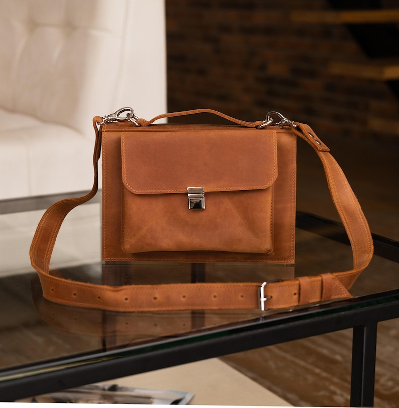 May include: A brown leather messenger bag with a flap closure and adjustable shoulder strap. The bag features a front pocket with a silver-colored clasp. The bag is sitting on a glass surface.