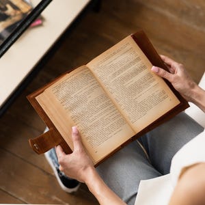 May include: An open book with aged pages is held in a brown leather cover. The book is being read, with the reader's hands visible. The book's pages are filled with text, and the cover has a button closure.