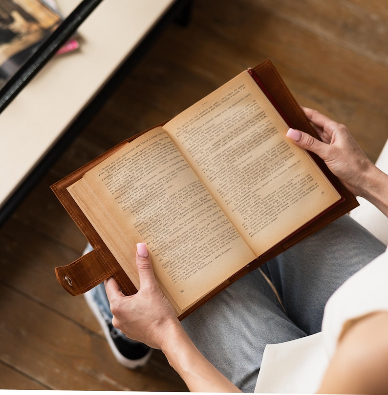 May include: An open book with aged pages is held in a brown leather cover. The book is being read, with the reader's hands visible. The book's pages are filled with text, and the cover has a button closure.