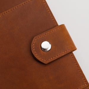 May include: Close-up of a brown leather journal with a silver snap closure. The leather has a textured appearance with visible stitching around the edges and on the closure flap. The journal appears to be new.