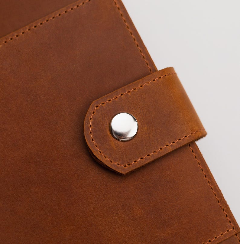 May include: Close-up of a brown leather journal with a silver snap closure. The leather has a textured appearance with visible stitching around the edges and on the closure flap. The journal appears to be new.
